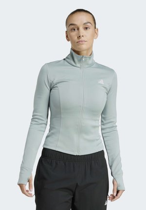 Mujer con una chaqueta deportiva gris claro con cremallera y agujeros para los pulgares, y pantalones deportivos negros, frente a un fondo liso.