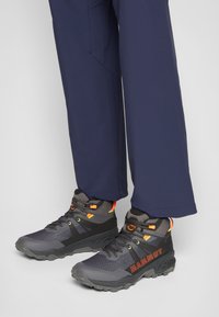 Încălțăminte de drumeție negre și gri cu accente portocalii, având un exterior texturat, vârf rotund și talpă din cauciuc. Purtați cu pantaloni bleumarin.