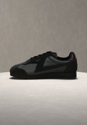 Baskets basses noires avec panneaux en daim et cuir, lacets à l'avant, et semelle en caoutchouc dentelée sur une surface claire.