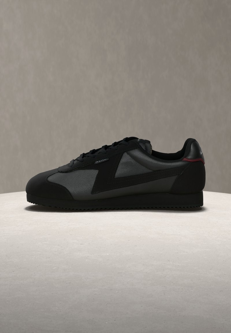 Μαύρο χαμηλό αθλητικό παπούτσι με πάνελ από σουέντ και δέρμα, μπροστινό δετό και οδοντωτή λαστιχένια σόλα σε ανοιχτό φόντο.