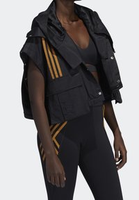 Černá zkrácená bunda s kapucí, zdobená oranžovými vertikálními pruhy, má více kapes a texturovanou látku. V kombinaci s černými legínami.