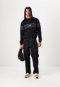 Sudadera negra con franjas grises y logo, combinada con pantalones negros con costuras en contraste. El modelo sostiene un pequeño bolso negro y lleva zapatillas beige.