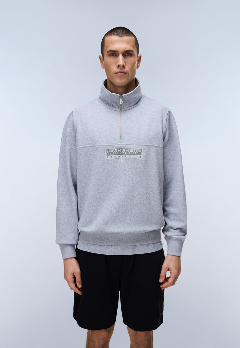 Νέος άνδρας που φοράει γκρι ανοιχτόφωμο φούτερ με φερμουάρ μέχρι το στήθος και λογότυπο, καθώς και μαύρο σορτς, στέκεται μπροστά σε απλό ανοιχτόφωμο φόντο.