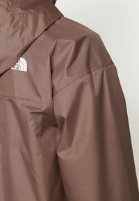Épaule et haut du dos d'une veste imperméable marron avec le logo blanc The North Face sur le côté gauche sous le col.