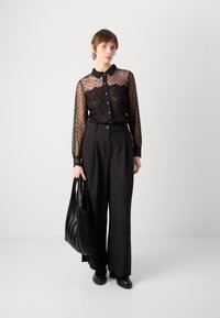 Blouse transparente noire à pois avec détails en dentelle, associée à un pantalon noir taille haute, portant un sac fourre-tout noir texturé et des chaussures noires.