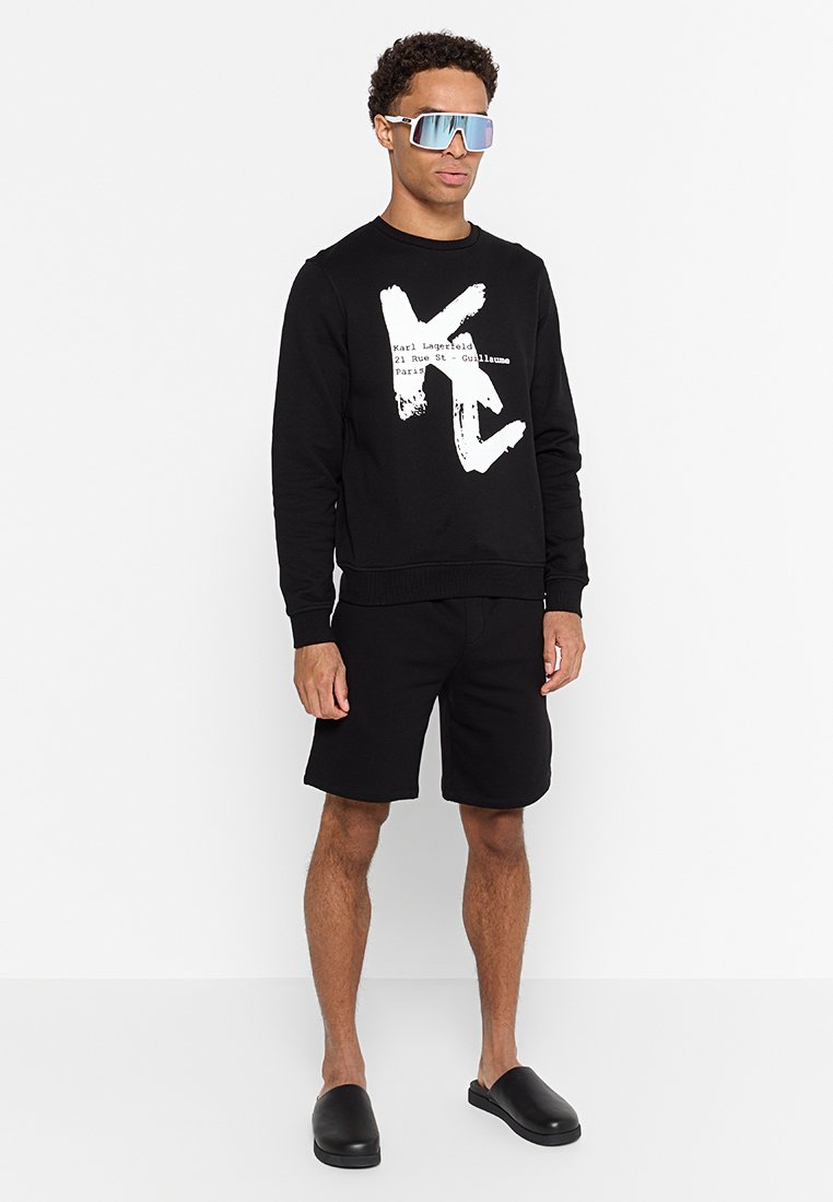 Sudadera negra con texto gráfico blanco; combinada con pantalones cortos negros. Gafas de sol elegantes con lentes espejados y zapatos negros de fácil colocación.