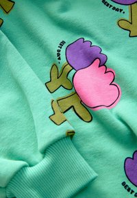 Tissu vert avec des fleurs de dessin animé colorées en rose et violet et le texte "BEST DAY" près des fleurs, avec des détails de poignets côtelés.