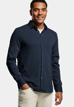 Chemise à manches longues navy boutonnière en tissu lisse, avec un col classique et des boutons contrastants, associée à un pantalon beige clair.
