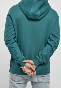 Sudadera con capucha de color verde azulado, mangas largas, tela con textura y puños acanalados, combinada con jeans azul claro. Vista trasera.