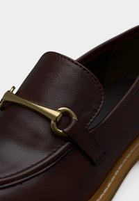 Σκούρο καφέ δερμάτινο μοκασίνι με χρυσή μεταλλική λεπτομέρεια αλογοουράς και ανοιχτό καφέ σόλα με λευκά ράμματα.