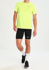 Camiseta deportiva amarilla neón de manga corta con un logo gris, combinada con pantalones cortos de compresión negros. Zapatillas negras y calcetines deportivos blancos completan el atuendo.