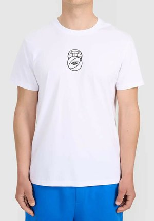 Mężczyzna w białej koszulce z krótkim rękawem z małym czarnym sportowym logo na piersi i jasnoniebieskich spodniach, stojący na jednolitym tle.