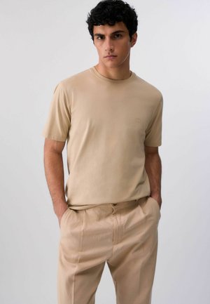 Giovane uomo con capelli ricci e scuri che indossa una t-shirt beige a maniche corte e pantaloni beige abbinati, con le mani nelle tasche, su uno sfondo semplice.