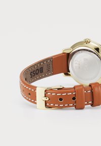 Correa de reloj de cuero marrón con costuras blancas, herrajes en tono dorado y caja redonda. La parte trasera tiene una tapa de cristal y detalles de marca.
