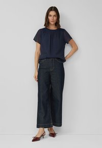 Frau steht vor einer weißen Wand und trägt eine marineblaue Bluse mit kurzen Ärmeln, dunkle Jeans mit weitem Bein und burgunderrote spitze High Heels.