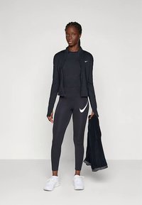 Black athletic outfit featuring a fitted long-sleeve jacket and leggings with a white swoosh logo, paired with white sneakers.