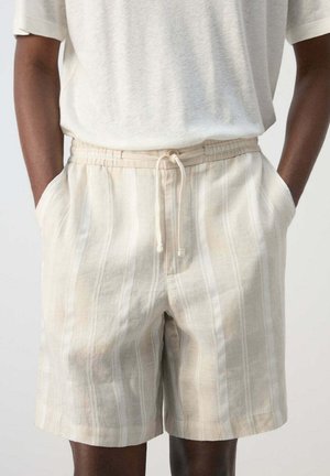 Hombre con pantalones cortos beige y blancos a rayas verticales con cordón y manos en los bolsillos, combinado con una camisa blanca de manga corta.