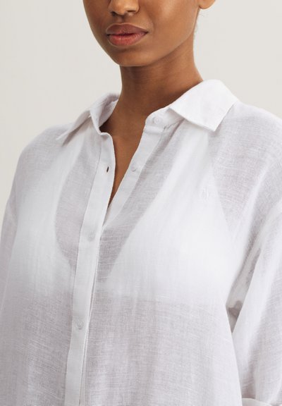 Λευκό λινό πουκάμισο με κουμπιά μπροστά, γιακά και ελαφρώς διάφανο ύφασμα, με διακριτικό λογότυπο πάνω από την αριστερή τσέπη.
