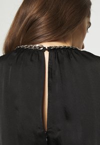 Femme aux longs cheveux bruns portant un haut noir texturé avec un détail de chaîne argentée au niveau de l'encolure arrière et une ouverture en forme de trou de serrure.