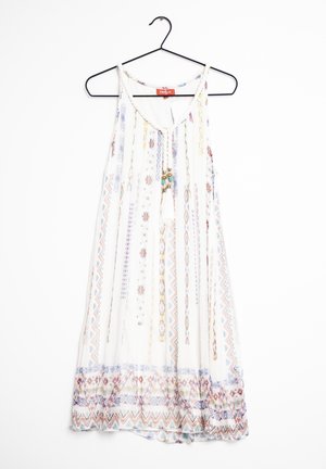 Hvid ærmeløs boho-kjole med farverige lodrette og kantede geometriske mønstre, kvastede snore, hængende på en sort bøjle.