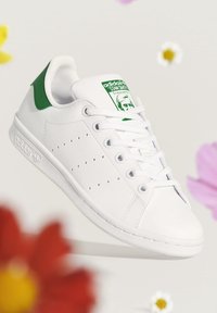 Bílé tenisky Adidas Stan Smith se zeleným patním okrajem, perforovanými bočními pruhy a značkovým jazykem, plovoucí na pozadí s rozmazanými barevnými květinami.