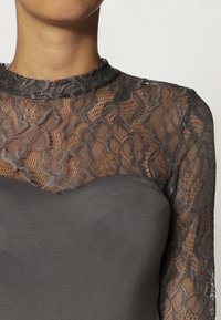 Grå klänning med en figursydd överdel och genomskinliga spetslångärmar. Spetsen har ett blommönster med vågiga kanter som ger textur och detaljer.