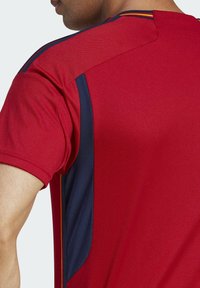Červené atletické tričko s texturovanou látkou, s tmavě modrými bočními panely a žlutými akcenty na ramenou.