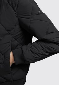 Čierna prešívaná bomber bunda s rebrovým manžetovým zakončením, bočnou kapsou a okrúhlym logom na rukáve, vyrobená z hladkého ľahkého materiálu.