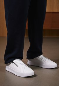 Hvite joggesko med en glatt lærfinish, flat såle og snøring. Sidelommer med glidelås gir funksjonalitet. Kombinert med mørke joggebukser.