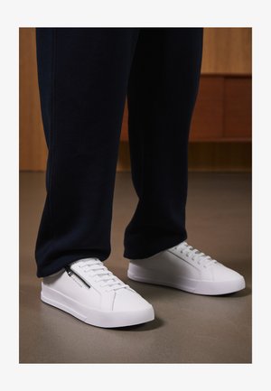 Weiße Sneaker aus glattem Leder, mit flacher Sohle und Schnürdesign. Details mit seitlichem Reißverschluss erhöhen die Funktionalität. Kombiniert mit dunklen Jogginghosen.