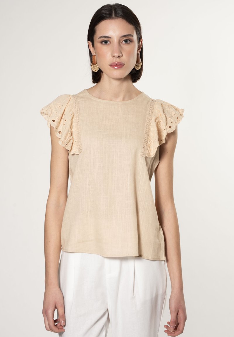 Top beige con maniche di pizzo a volant e delicati ricami lungo le spalle. Realizzato in tessuto leggero, abbinato a pantaloni bianchi.