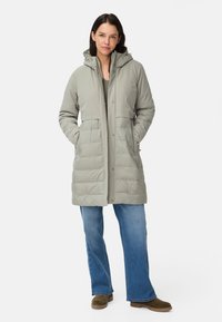 Manteau matelassé gris clair, longueur genou, avec capuche, manches longues, deux poches avant et une texture lisse. Associé à un jean évasé bleu et des bottes marron.