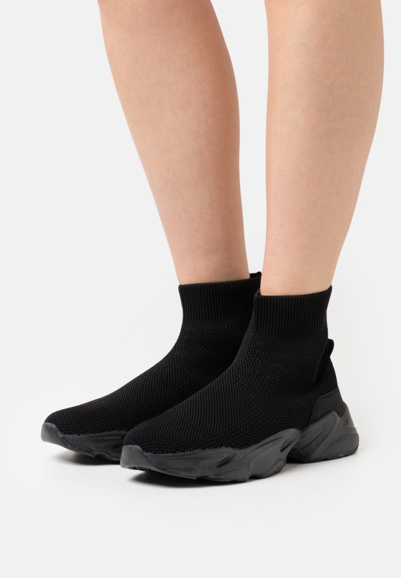 Jambes portant des baskets style chaussettes tricotées noires avec des semelles épaisses texturées, présentées sur un fond blanc uni.