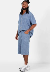 Camisa de manga corta azul claro con cierre de botones, que presenta un patrón sutil, combinada con pantalones cortos a juego. Usada con zapatillas deportivas plateadas.