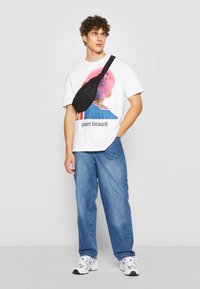 Hvit grafisk t-skjorte med et fargerikt design som har en figur og tekst. Kombinert med lyseblå jeans og en svart veske over skulderen. Sneakersene er hvite.