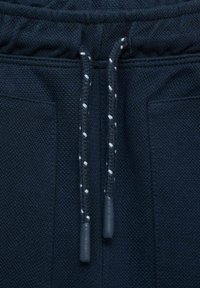 Detailní záběr na tmavě modré pletené kalhoty s elastickým pasem, které mají stahovací šňůrku s bílými akcenty a boční kapsy.