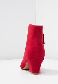 Czerwone zamszowe botki za kostkę z tępo zakończonym obcasem, gładką teksturą i małym detalem w postaci kokardy z tyłu. Czyste linie i minimalistyczny design.