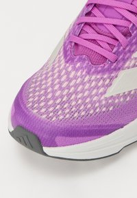 Kevyt juoksukenkä, jossa on violetti verkkomateriaali yläosa, teksturoitu muotoilu, vahvistettu varvasosa ja harmaa ulkopohja valkoisilla yksityiskohdilla.