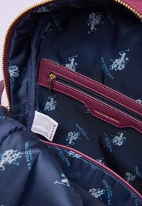 Interiör av en väska med marinblått tyg och upprepade ljusblå US Polo Assn. logotyper. Inkluderar en vinröd zipficka och vårdlabel.