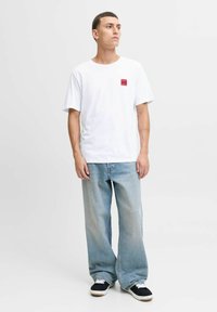 Junger Mann steht, trägt ein weißes T-Shirt mit rotem Logo, lockere hellblaue Jeans und schwarze Sneaker mit weißen Schnürsenkeln vor einem einfarbigen Hintergrund.