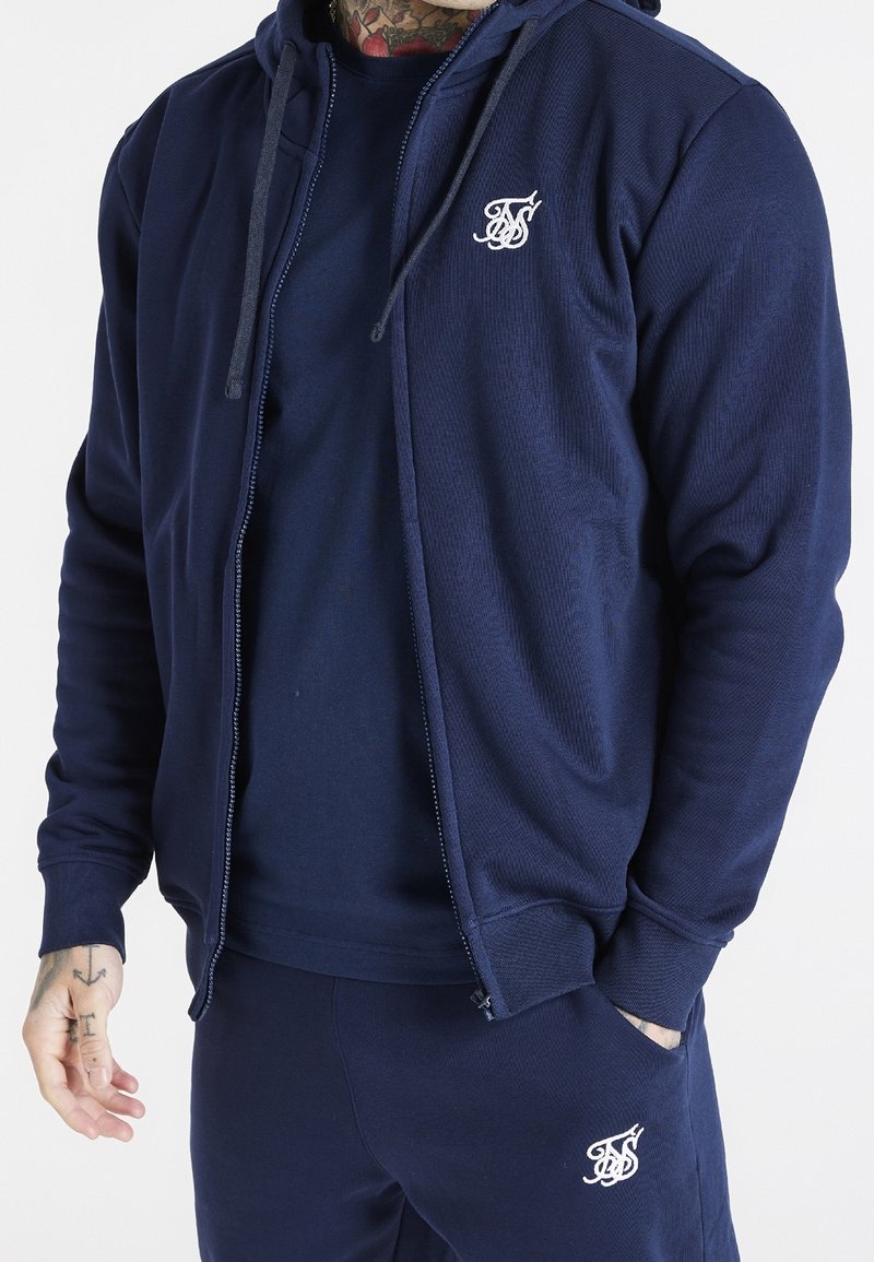 Homme portant un sweat à capuche bleu marine zippé, un pantalon assorti et un t-shirt bleu marine, tous avec des logos brodés blancs sur la poitrine et la cuisse.