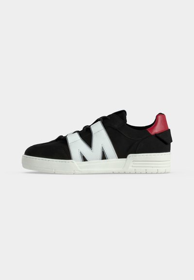 Μαύρο sneaker χαμηλής κοπής με παχύ λευκό πέλμα, μεγάλο λευκό γράμμα Μ στο πλάι και κόκκινη λεπτομέρεια στη φτέρνα πάνω σε απλό ανοιχτό φόντο.