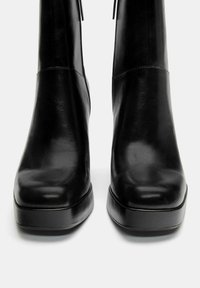 Crne kožne čizme do gležnja s glatkom teksturom, kvadratnim vrhom i debelim potplatom. Imaju minimalno šivanje i elegantan, moderni dizajn.