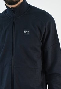 Chaqueta de cremallera azul marino hecha de tela suave; cuenta con un cuello alzado, bolsillos laterales y un logo de EA7 en el pecho.