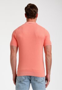 Trič eo s krátkým rukávem v korálové barvě, s texturovaným žebrovaným vzorem a klasickým kulatým výstřihem, nošené s světlými modrými džíny.