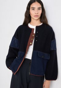 Giacca in misto lana blu navy con accenti in denim, chiusure a scatto e zip, due tasche frontali e colletto rotondo. Presenta dettagli a contrasto in marrone.