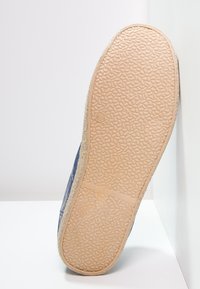 Semelle d'une chaussure en tissu bleu avec une semelle en caoutchouc beige texturée indiquant la taille 41, reposant verticalement contre un coin blanc.