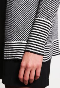 Schwarz-weiß gestreifte Strickjacke mit strukturiertem Strickmuster und einfarbigem schwarzen Saum. Die Ärmelsäume haben abwechselnde horizontale Streifen.