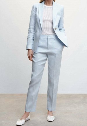 Kobieta w jasnoniebieskim, dopasowanym garniturze z marynarką na miarę, dopasowanymi spodniami, białym prążkowanym topem i białymi balerinami stoi na szarej podłodze.