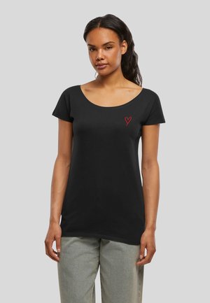 Femme aux cheveux foncés portant un t-shirt noir à col rond avec un petit cœur rouge sur la poitrine gauche et un pantalon gris clair.
